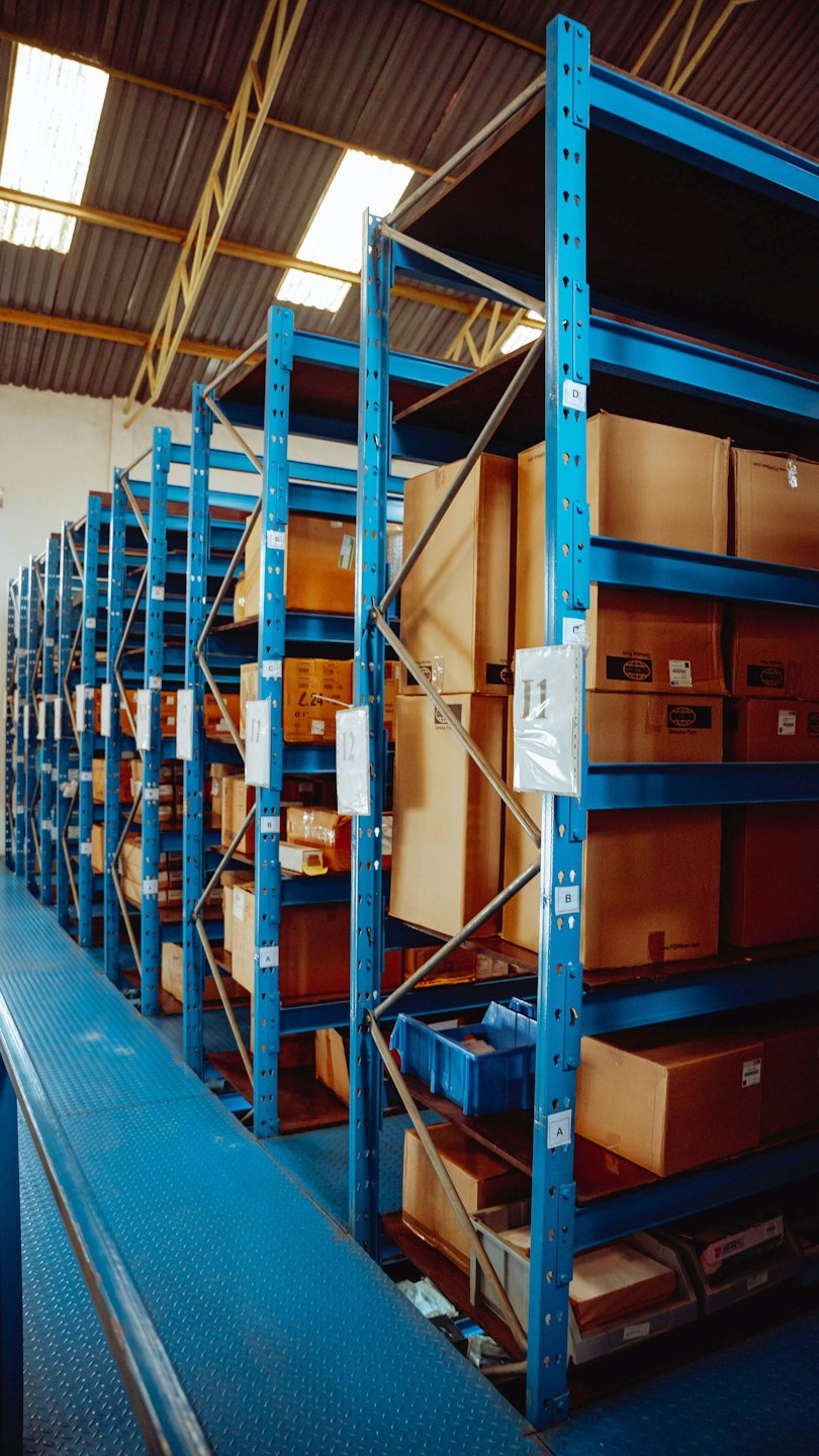 Pasillo de almacén con estanterías altas y azules apiladas con cajas.